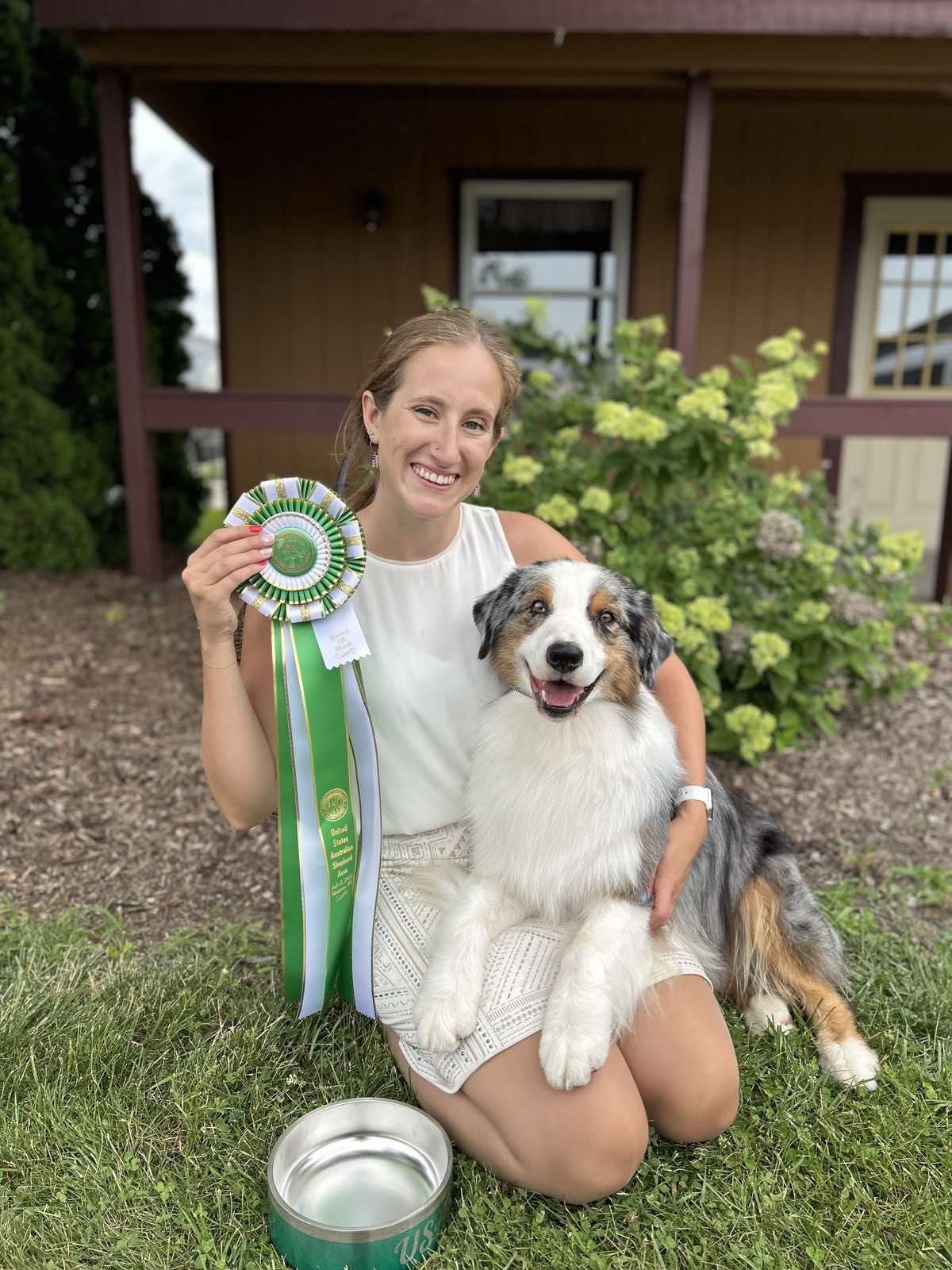 Blue merle Australian Shepherd 'Ruzek' (AKC CH Thornapple Roger That) sitting with his handler and a United States Australian Shepherd Association rosette